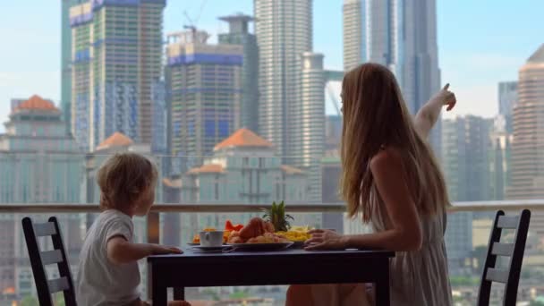 Jonge vrouw gouvernante ontbijten met een jongen op een balkon met uitzicht op de wolkenkrabbers van het centrum van de stad — Stockvideo