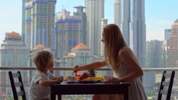 Jonge vrouw gouvernante ontbijten met een jongen op een balkon met uitzicht op de wolkenkrabbers van het centrum van de stad — Stockvideo