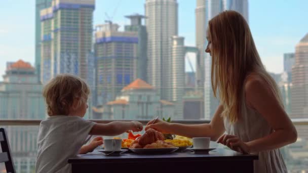 Jovem governanta tomar café da manhã com um menino em uma varanda com vista para os arranha-céus do centro da cidade — Vídeo de Stock