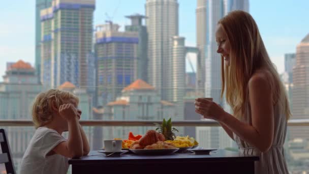 若い女性の知事は男の子と市内中心部の高層ビルを見渡すバルコニーで朝食を持っています。 — ストック動画
