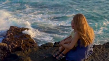 Bir anne ve oğlu bir okyanus dalgalarının yakın Tanah Lot tapınağın bakarak bir kayanın üzerine oturmuş Slowmotion atış