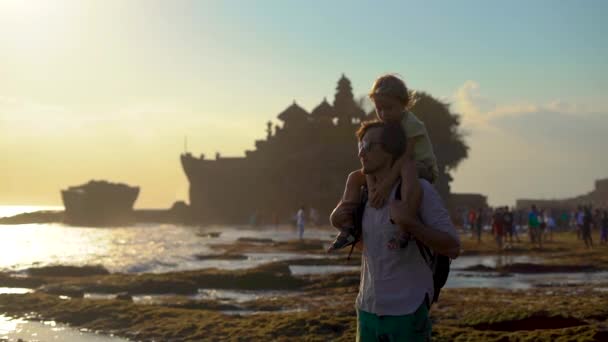 Zeitlupenaufnahme eines Vaters und seines Sohnes, die bei Sonnenuntergang an einer Küste in einem wunderschönen Tempel auf der Bali-Insel spazieren — Stockvideo