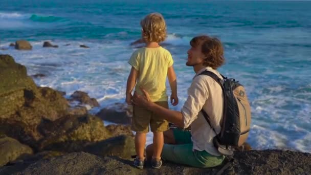 Slowmotion lövés egy Atya és a fiú ült egy sziklán, látszó-on egy óceán hullámai a Tanah Lot-templom a közelben — Stock videók