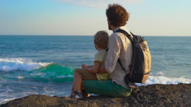 Медленный снимок отца и сына, сидящих на скале и смотрящих на океанские волны рядом с храмом Тана Лота — стоковое видео