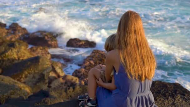 Fotografía en cámara lenta de una madre y un hijo sentados en una roca mirando las olas del océano cerca del templo de Tanah Lot — Vídeos de Stock