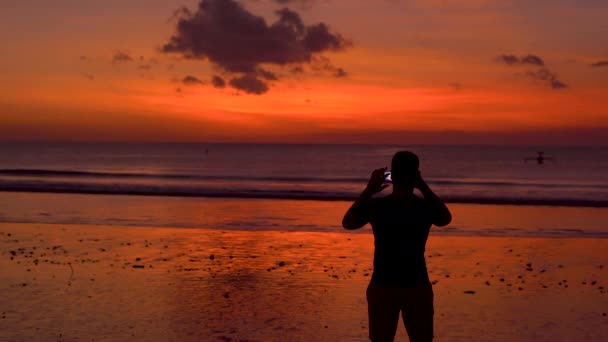 浜辺で美しい日没時に彼の携帯電話で写真を撮る人のシルエット — ストック動画