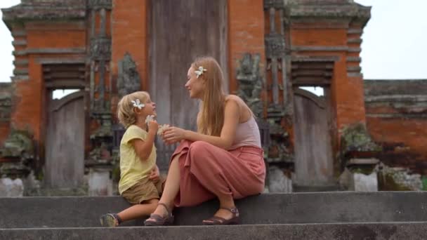 Handhållna skott av en ung kvinna och hennes son som sitter på trappan till Taman Ayun templet på ön Bali. — Stockvideo