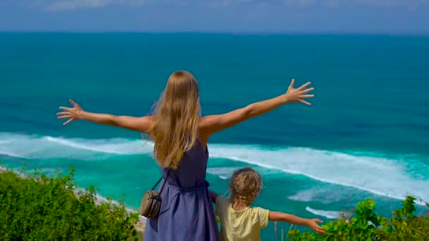 Slowmotion shot of a young woman and her son standing at the edge of the cliff looking at the ocean. Suluban and Nyang Nyang place. Traveling with kids concept — Stock Video