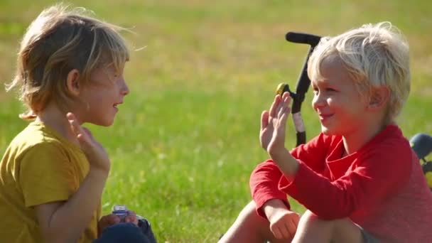 Zeitlupenaufnahme von zwei kleinen Jungen Radler sitzen auf einer Wiese und geben einander fünf. Erfolgskonzept. Freundschaftskonzept. — Stockvideo