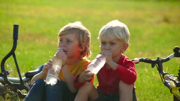 Slow Motion shot av två små pojkar cyclers sitta på en gräsmatta och dricka vatten från en plastflaskor. Sötvatten koncept. Vänskap-konceptet. — Stockvideo