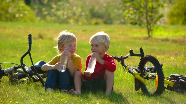 Slow Motion shot of two little boys cyclers sit on a lawn and drink water from a plastic bottles. Freshwater concept. Friendship concept. — Stock Video