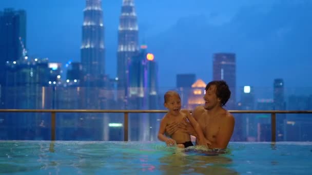 Tikje shot van een jonge man en zijn zoontje met plezier in een zwembad op het dak met uitzicht op de wolkenkrabbers — Stockvideo