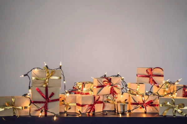 Pile of gifts wrapped in craft paper with red and gold ribbons on black wooden table with fairy lights for Christmas holiday.