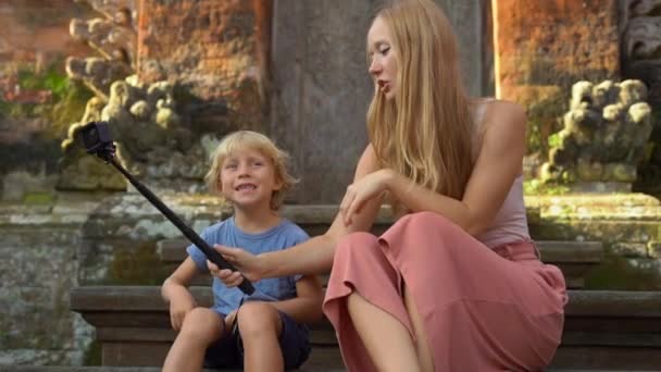 Fotografía en cámara lenta de una hermosa joven y su pequeño hijo hacen una selfie con una cámara de acción en las escaleras del templo balinés. Viajar a Indonesia concepto. Viajar con concepto de niños — Vídeos de Stock