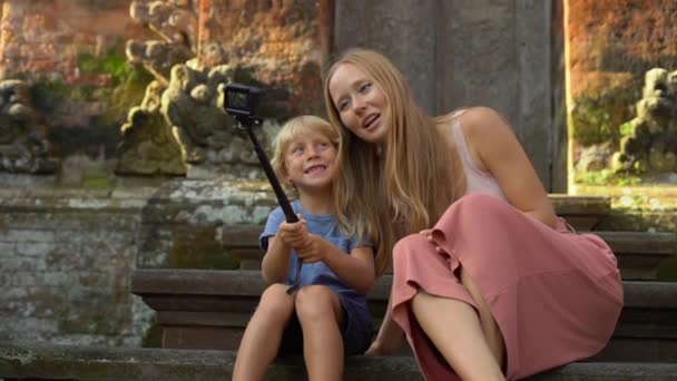 Fotografía en cámara lenta de una hermosa joven y su pequeño hijo hacen una selfie con una cámara de acción en las escaleras del templo balinés. Viajar a Indonesia concepto. Viajar con concepto de niños — Vídeos de Stock