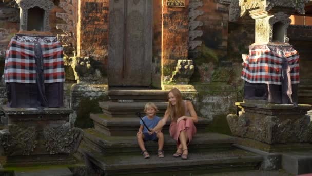 一个美丽的年轻女子和她的小儿子在巴厘岛寺庙的楼梯上用动作相机做了一个自拍 前往印度尼西亚的概念 带孩子旅行的概念 — 图库视频影像