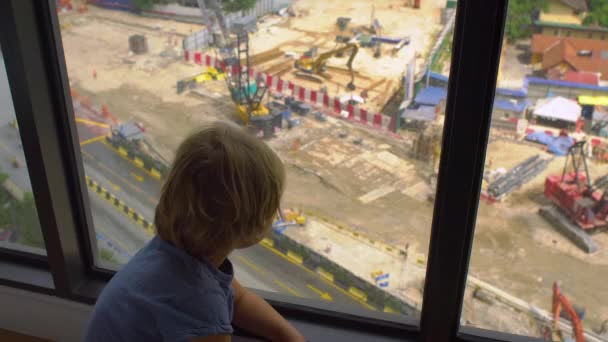 Niño pequeño a través de la ventana mirando un sitio de construcción de un edificio alto — Vídeos de Stock