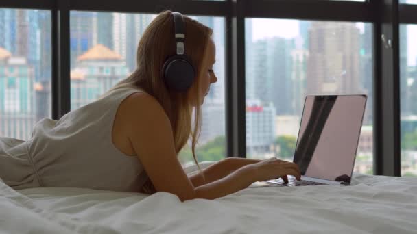 Молодая женщина-фрилансер, лежащая на кровати, слушает музыку с помощью беспроводных наушников и работает над своим ноутбуком — стоковое видео
