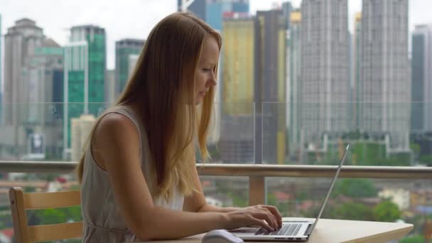 Mladá žena na volné noze pracoval na svém laptopu na balkón s pozadím města centrum plné mrakodrapů. — Stock video