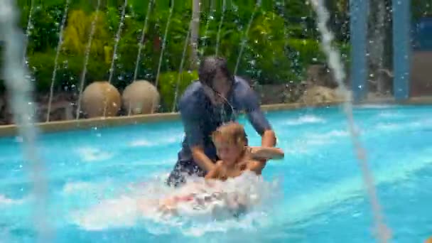 Медленный снимок молодого отца и его сына, веселящихся в тропическом бассейне среди фонтанов — стоковое видео