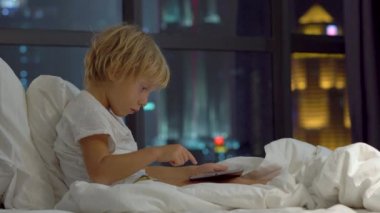 küçük çocuk geceleri yatağında oturup bir gökdelenin siluetleri ile tablet pc bir arka plan çalış..