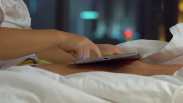 Primer plano de un niño sentado en su cama por la noche toca una tableta PC con siluetas de un rascacielos en un fondo — Vídeo de stock
