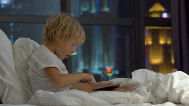 Niño sentado en su cama por la noche juega una tableta PC con siluetas de un rascacielos en un fondo — Vídeos de Stock