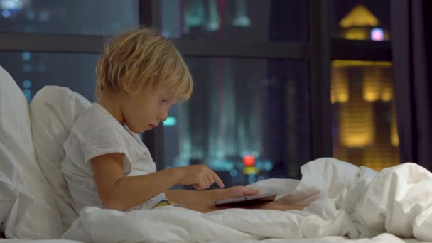晚上坐在床上的小男孩在后台玩平板电脑 有摩天大楼的剪影 — 图库视频影像