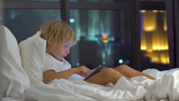 Menino sentado em sua cama à noite desempenha um PC tablet com silhuetas de um arranha-céu em um fundo — Vídeo de Stock