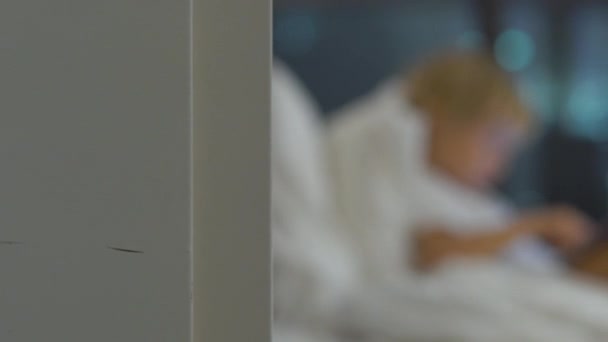 Jongetje in zijn bed zitten 's nachts speelt een tablet pc met silhouetten van een wolkenkrabber bij een achtergrond — Stockvideo