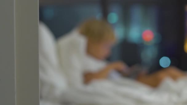 Kisfiú ül az ágyában, éjjel a háttérben játszik egy tabletta pc-val sziluettek egy felhőkarcoló — Stock videók