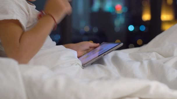 Küçük çocuk geceleri yatağında oturmuş portre çekim tablet pc bir gökdelenin siluetleri ile bir arka plan çalış. Mobil cihazlar bağımlılık kavramı. Çocuk eğitim kavramı. — Stok video