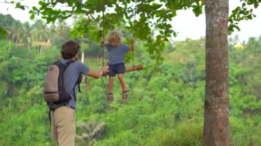 Genç adam küçük oğlu tropikal parkta salıncak iterek yavaş çekim. 