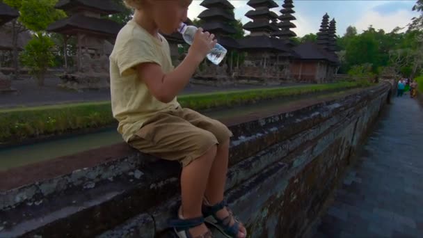 Ένα μικρό αγόρι που πίνει νερό από ένα πλαστικό μπουκάλι που κάθεται σε ένα τοίχο του ναού Taman Ayun πυροβολισμό slowmotion. Έννοια του καθαρού νερού — Αρχείο Βίντεο