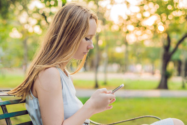 Young calm girl using her mobile phone to send a text while standing in a park