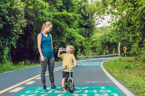 Mulher desportiva a correr na estrada ao nascer do sol. O filho vai equilibrar a bicicleta. Fitness e conceito de bem-estar de treino — Fotografia de Stock