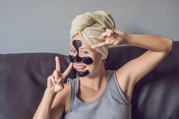 Güzel genç kadın evde yüz maskesi ile rahatlatıcı. Yüzünde siyah maske uygulayarak mutlu neşeli kadın — Stok fotoğraf