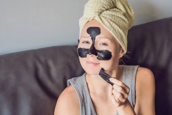 Hermosa mujer joven que se relaja con mascarilla en casa. Feliz mujer alegre aplicando máscara negra en la cara — Foto de Stock