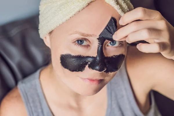 Mulher bonita relaxante com máscara facial em casa. Feliz mulher alegre aplicando máscara preta no rosto — Fotografia de Stock