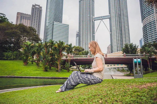 Eine junge Frau in Freizeitkleidung mit Laptop in einem tropischen Park vor dem Hintergrund von Wolkenkratzern. Mobiles Bürokonzept — Stockfoto