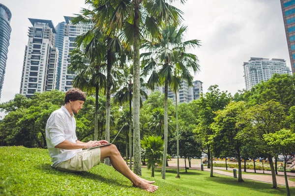 Férfi üzletember vagy diák alkalmi ruha segítségével laptop egy trópusi parkban a háttérben felhőkarcolók. Fehér ingbe öltözve, bézs rövidnadrágban. Mobil iroda koncepciója — Stock Fotó