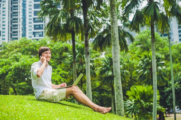 Mann Geschäftsmann oder Student in Freizeitkleidung mit Laptop in einem tropischen Park vor dem Hintergrund von Wolkenkratzern. Bekleidet mit einem weißen Hemd, beigen Shorts. Mobiles Bürokonzept — Stockfoto