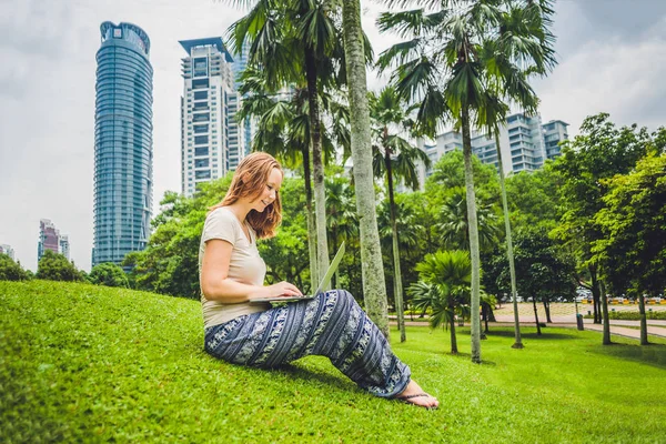 Eine junge Frau in Freizeitkleidung mit Laptop in einem tropischen Park vor dem Hintergrund von Wolkenkratzern. Mobiles Bürokonzept — Stockfoto