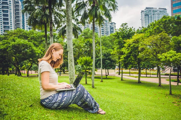 캐주얼 드레스를 입은 한 젊은 여성이고 층 건물의 배경에 있는 열대 공원에서 노트북을 사용하고 있습니다. 모바일 오피스 개념 — 스톡 사진