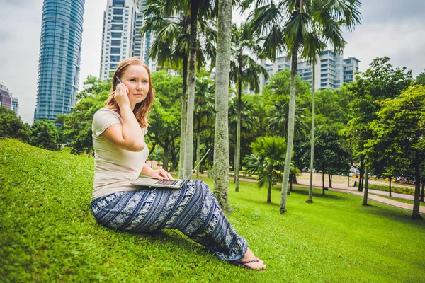 Une jeune femme en tenue décontractée utilisant un ordinateur portable dans un parc tropical sur fond de gratte-ciel. Concept de bureau mobile — Photo