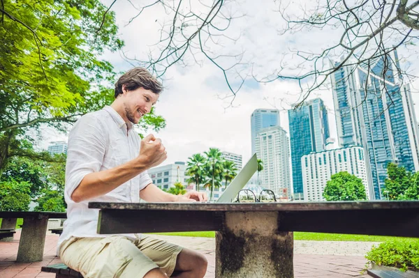 在热带公园的摩天大楼背景下，穿着休闲装的商人或学生正在使用笔记本电脑。穿着一件白色衬衫，米黄色短裤。流动办公室概念 — 图库照片