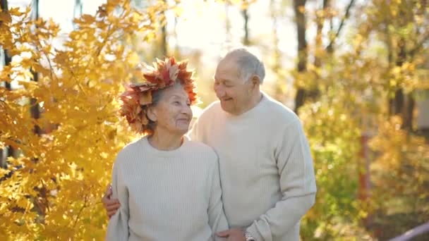 Kieliszek w slowmotion pary staruszków przytulanie i uśmiechając się do siebie w parku w przepięknym otoczeniu jesień. Stara kobieta nosi wieniec z liści jesienią — Wideo stockowe