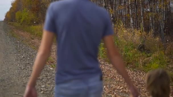 Fotografía en cámara lenta de un padre y su hijo caminando por la carretera con un bosque de otoño cerca y muchas hojas amarillas cayendo al suelo. Concepto de viaje de otoño — Vídeos de Stock
