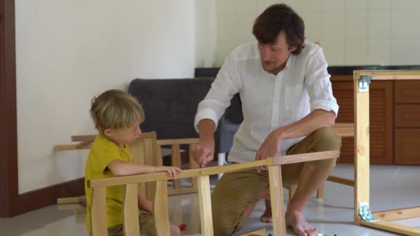 Vater und Sohn montieren Holzmöbel aus Kleinteilen. Kleiner Junge hilft seinem Vater, einen Stuhl zusammenzubauen. — Stockvideo