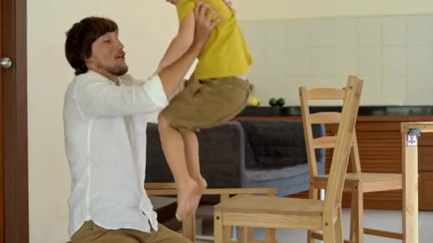 Vater und Sohn montieren Holzmöbel aus Kleinteilen. nach Beendigung einer Stuhlmontage setzt Mann seinen Sohn auf einen Stuhl und gibt ihm eine Fünf — Stockvideo